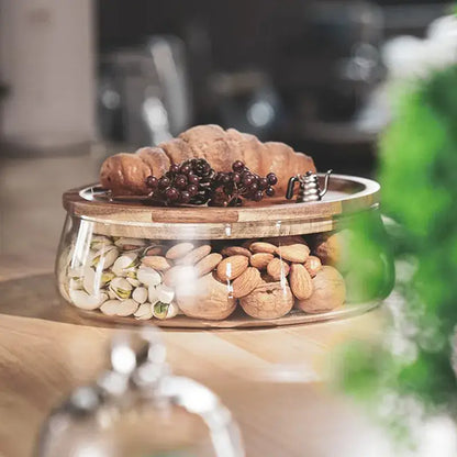 Glass food storage containers with wooden lids filled with mixed nuts topped with croissant grapes and a silver teapot