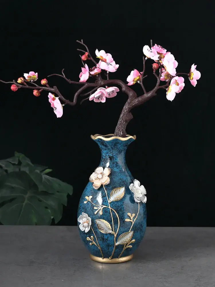 A blue ceramic vase with gold accents and floral embellishments holds dried flowers on a wobble plate display