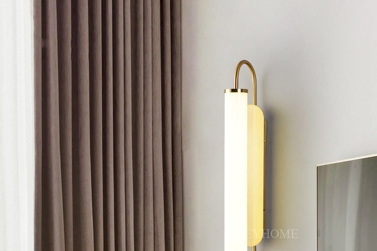 A sleek modern minimalist gold led wall lamp with a curved brass finish and a cylindrical frosted glass shade emitting warm light for bedroom use.