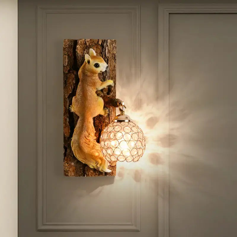 A nordic vintage style wall lamp featuring a charming golden resin squirrel with a textured tree trunk base and an ornate crystal-encrusted globe lampshade for bedroom lighting.