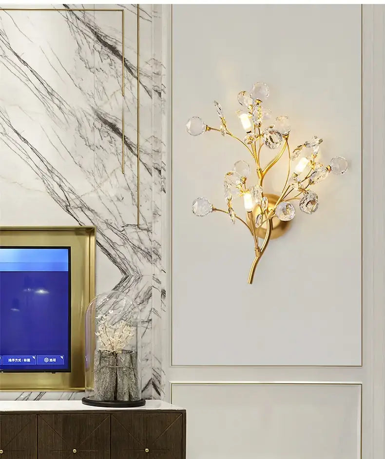 Post modern luxury gold-toned wall sconce featuring crystal branches and spherical accents designed as a luxury branch style bedroom light.