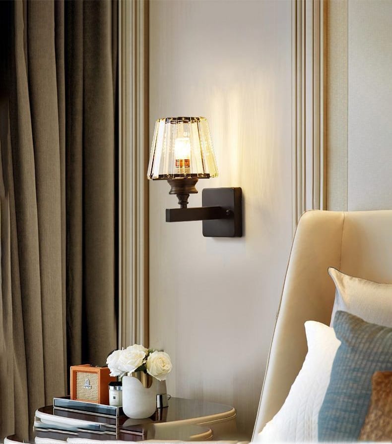 A warm-toned modern minimalist wall sconce featuring a faceted crystal glass shade and a dark metal arm designed as a minimalist wall lamp for bedroom use.