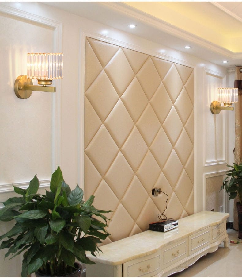 Beige quilted leather accent wall panel with diamond-shaped tufting behind a modern minimalist gold wall lamp with crystal shade for bedroom.