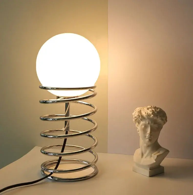 A sleek chrome bauhaus spring table lamp with a glowing white glass globe suitable as a bedroom light.