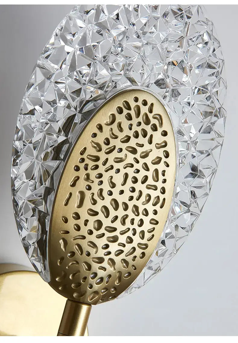 A modern gold-toned led wall lamp with an oval metal backplate featuring intricate droplet cut-outs and a faceted clear crystal circular frame, ideal for indoor lighting in the bedroom.