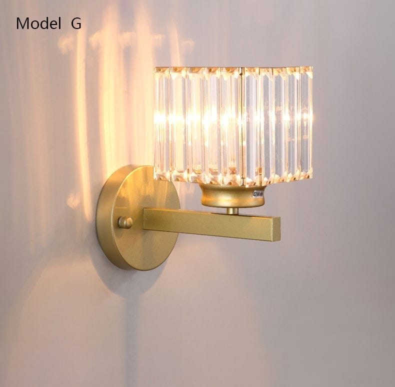 Modern minimalist wall sconce with a gold finish and a cylindrical crystal shade made of vertical faceted glass rods designed for bedroom use.