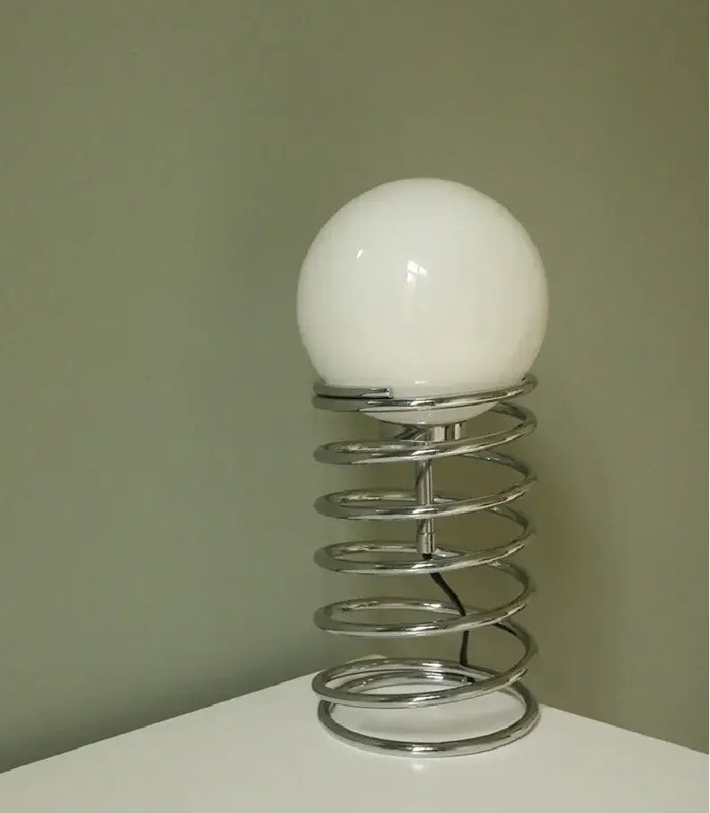 a bauhaus spring table lamp featuring a sleek chrome spring base and a smooth opaque white glass spherical lampshade designed for bedroom lighting