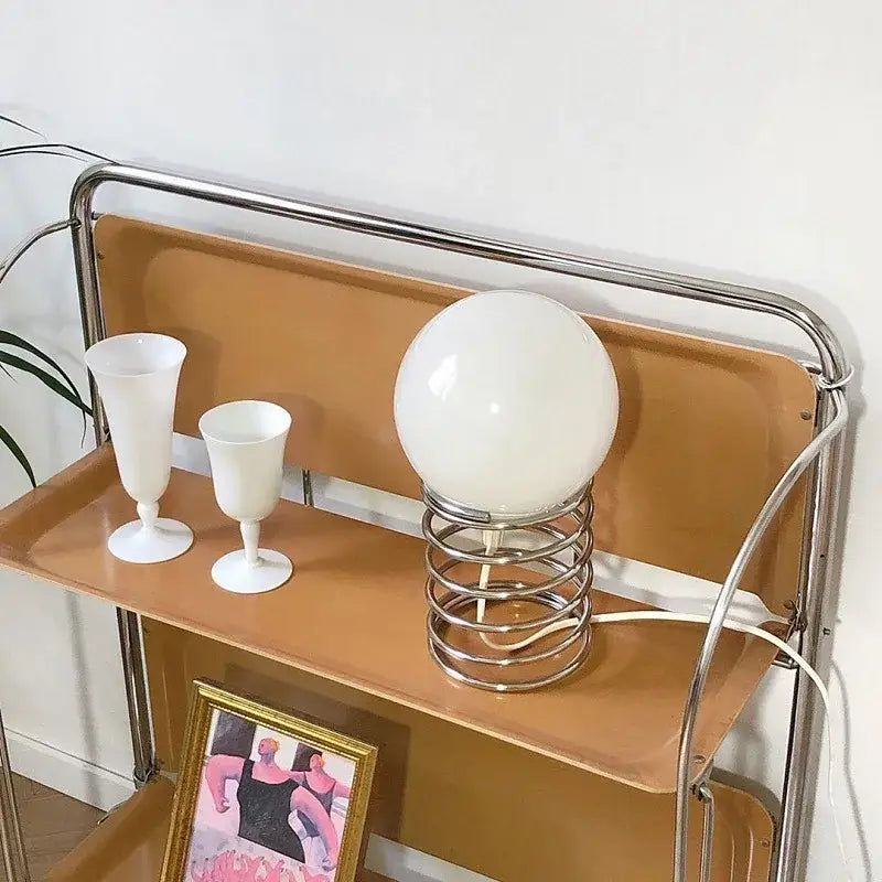 A sleek spherical white glass bauhaus spring table lamp with a chrome spring base sits elegantly on a wooden shelf as a bedroom light.