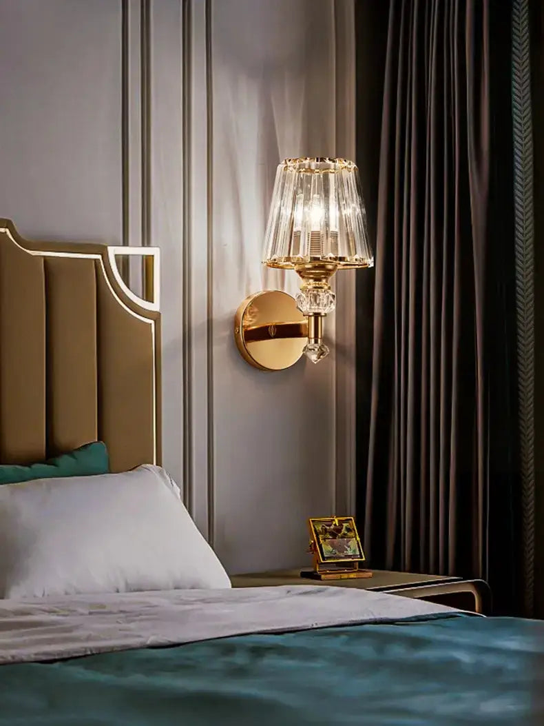 Modern minimalist wall sconce in gold tone featuring a faceted crystal shade and decorative crystal accents for bedroom lighting.