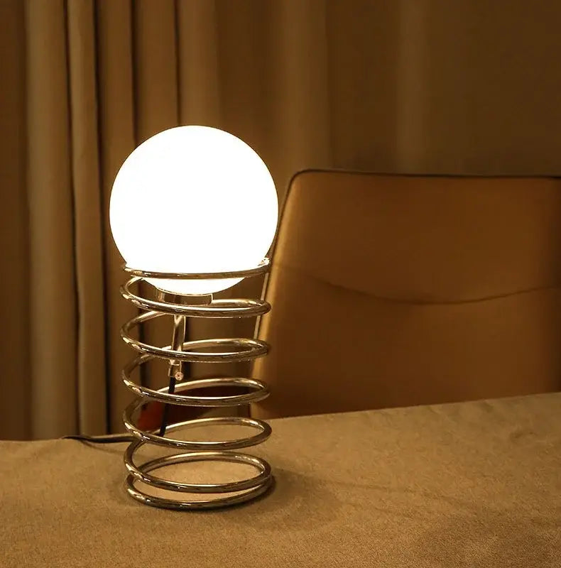 A bauhaus spring table lamp with a glowing white glass spherical bulb and a polished chrome spiral spring base ideal for bedroom lighting.
