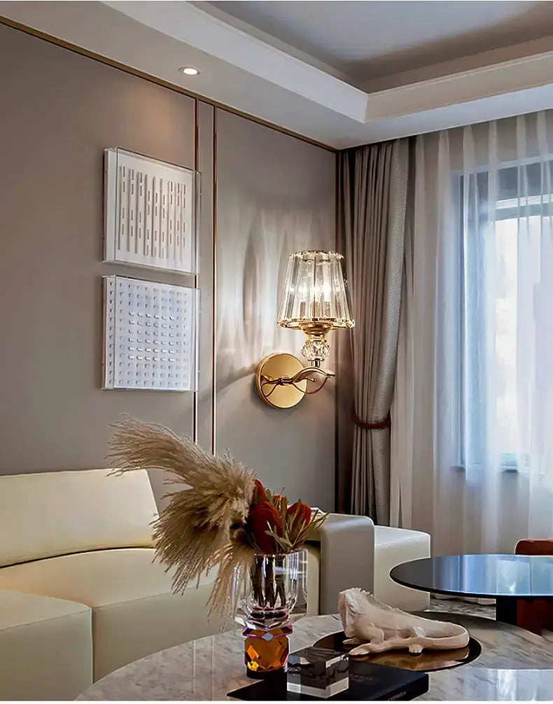 A modern minimalist gold wall lamp with a crystal shade illuminating a clear crystal vase holding dried pampas grass and red flowers in a bedroom setting.
