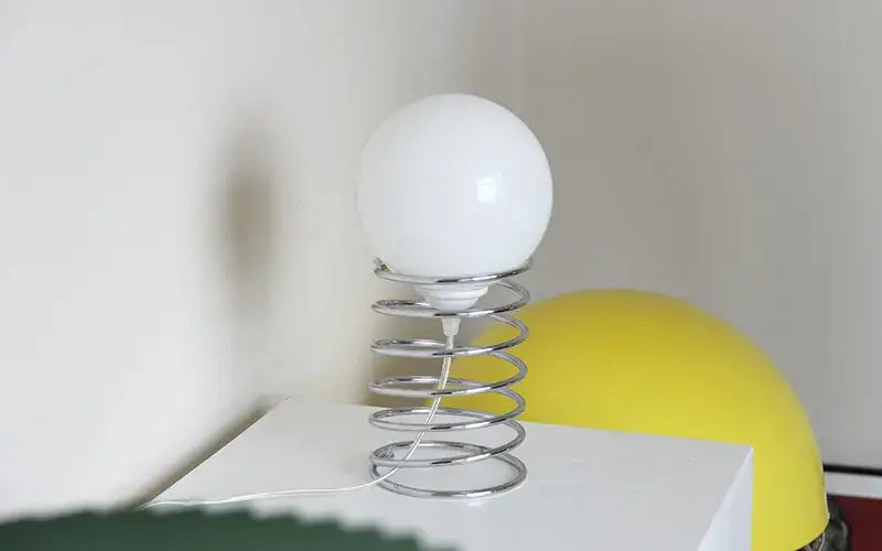 A sleek white glass bauhaus spring table lamp with a chrome spring base designed for bedroom lighting.
