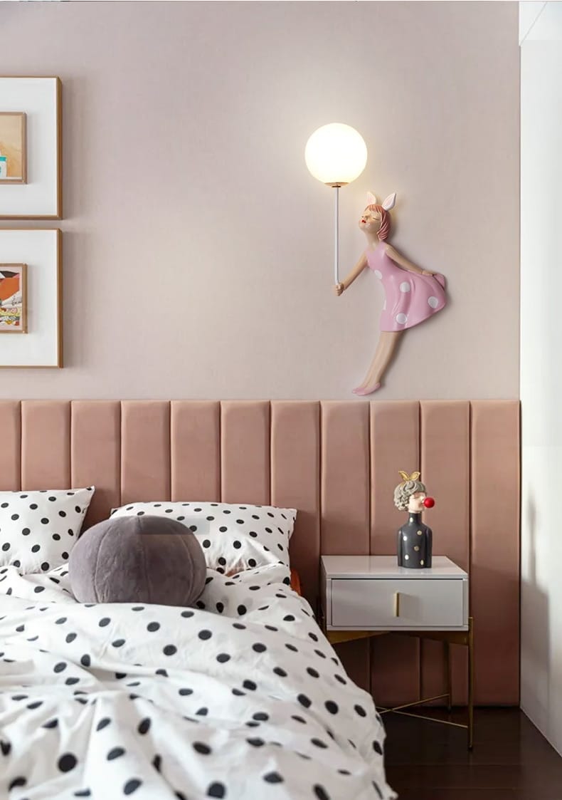 A whimsical pink polka dot fairy girl wall lamp featuring a pink fairy figurine wall sconce with a round white globe light for bedroom use.