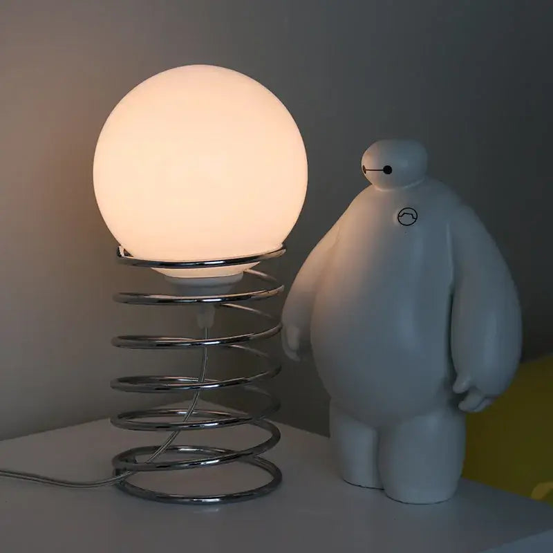 a bauhaus spring table lamp featuring a glowing white glass spherical shade and a chrome spring base designed as a bedroom light