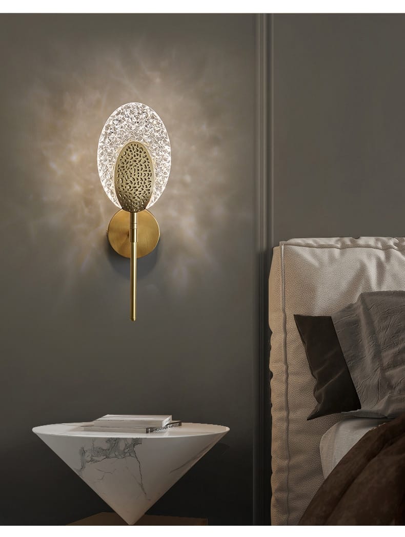 Gold-toned led wall lamp featuring a textured oval-shaped glass diffuser and an intricate patterned central element for indoor lighting in the bedroom.
