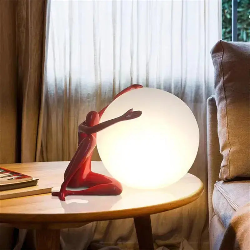 A sleek glossy red ball holding character resin figurine beside a glowing white lamp on a wooden table for living room