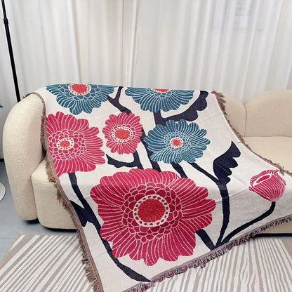 A cozy cream-colored throw blanket with bold pink and teal floral pattern and fringed edges featuring geometric knitted tassels
