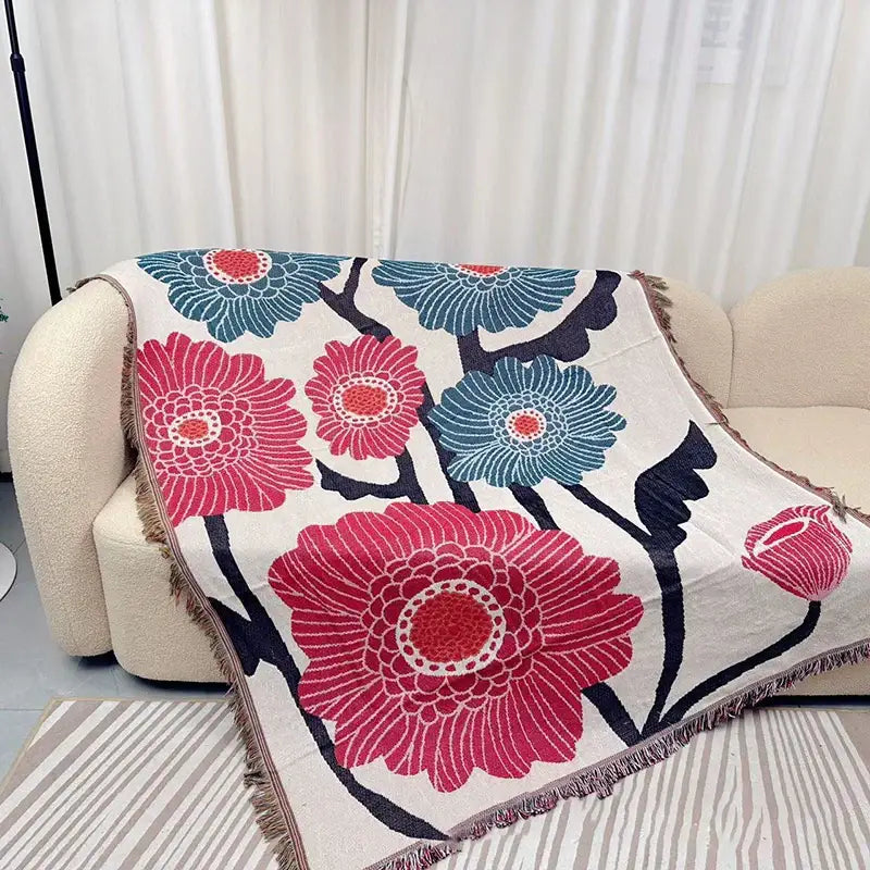 A cozy cream-colored throw blanket with bold pink and teal floral pattern and fringed edges featuring geometric knitted tassels
