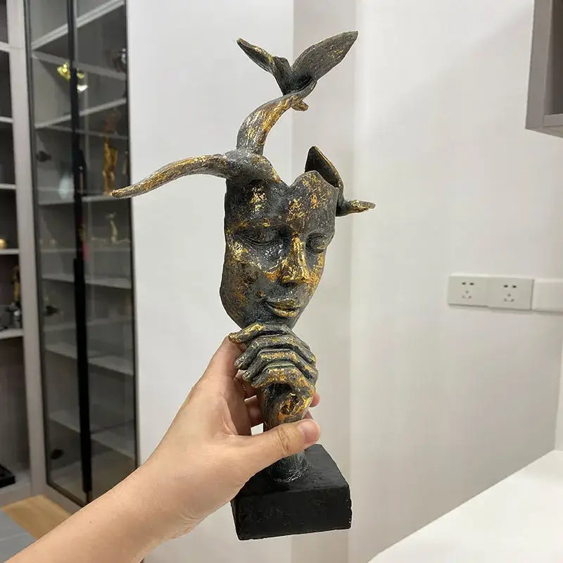 Abstract bronze and gold mask bird figurine statue featuring a human face with horns and a bird perched on the horn held in hand