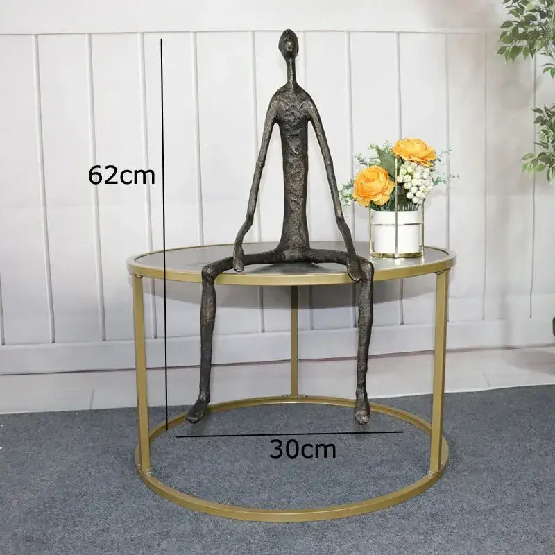 A dark bronze abstract metal human sculpture with elongated limbs seated on a gold-framed table for decorbites abstract metal decor