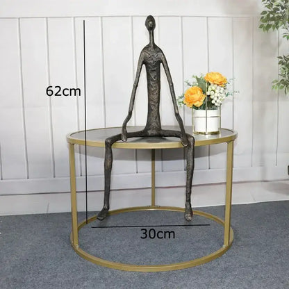 A dark bronze abstract metal human sculpture with elongated limbs seated on a gold-framed table for decorbites abstract metal decor