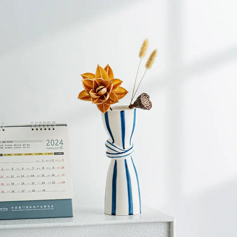 Abstract ceramic hydroponic vase in white ceramic with blue stripes and knotted design holding orange dried flowers