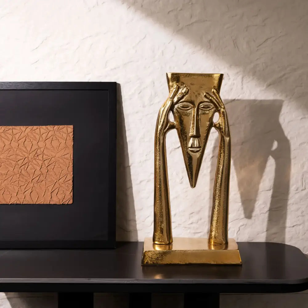 Abstract golden masked figure sculpture with elongated features and hands framing the head on square base for living room decor