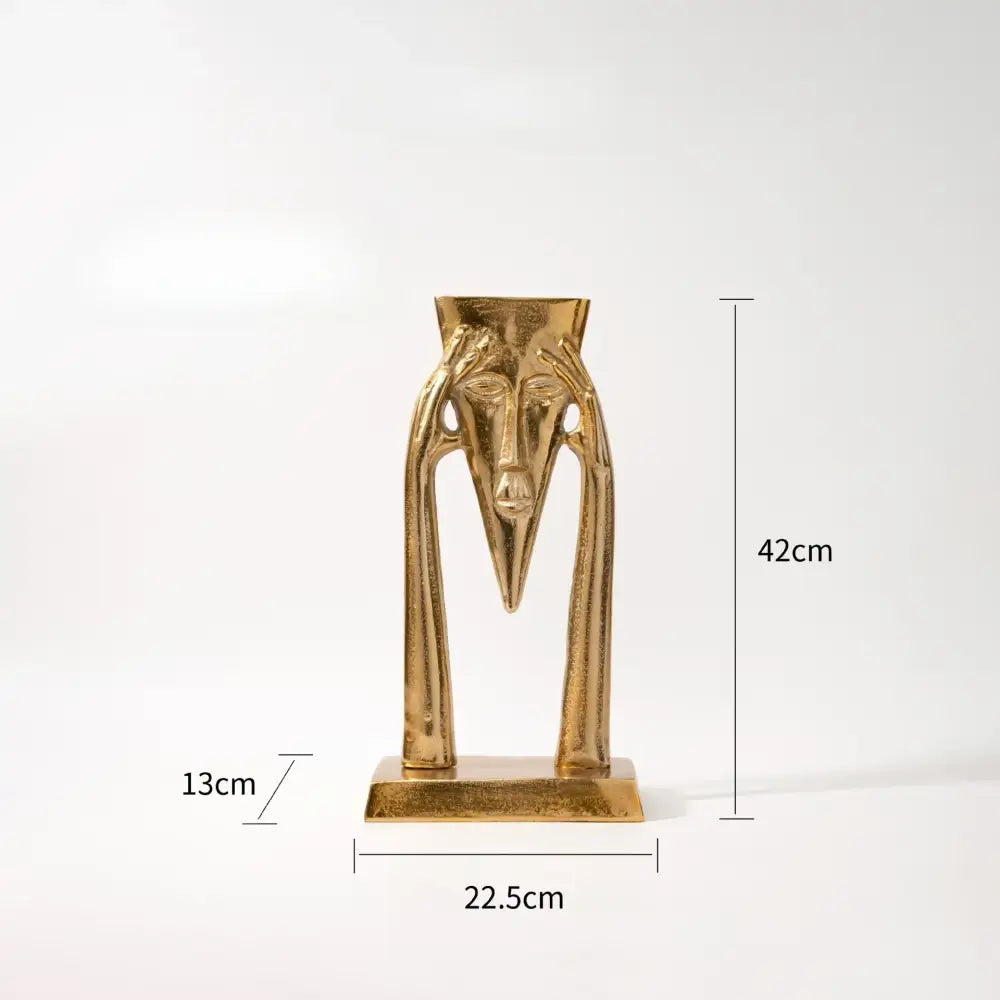 Golden metallic abstract masked figure sculpture with elongated face and curved arms for living room decor on rectangular base