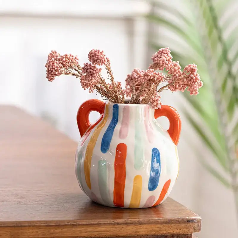 Colorful cartoon ceramic vase teapot with orange handles and multicolor stripes holding dried pink flowers for decorbites decor