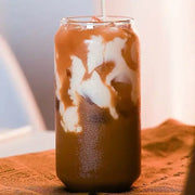 A tall clear glass from an aesthetic drinking glasses set filled with iced coffee and cream swirls with a lid and straw