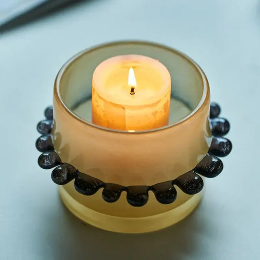 A translucent amber glass cylinder vase with black beaded rim holds a lit orange candle for living room decor