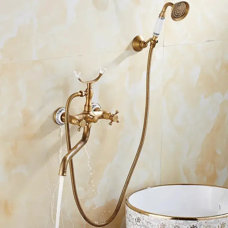 Antique brass bathtub shower set with ornate ceramic handles mounted on beige marble wall beside white vessel sink