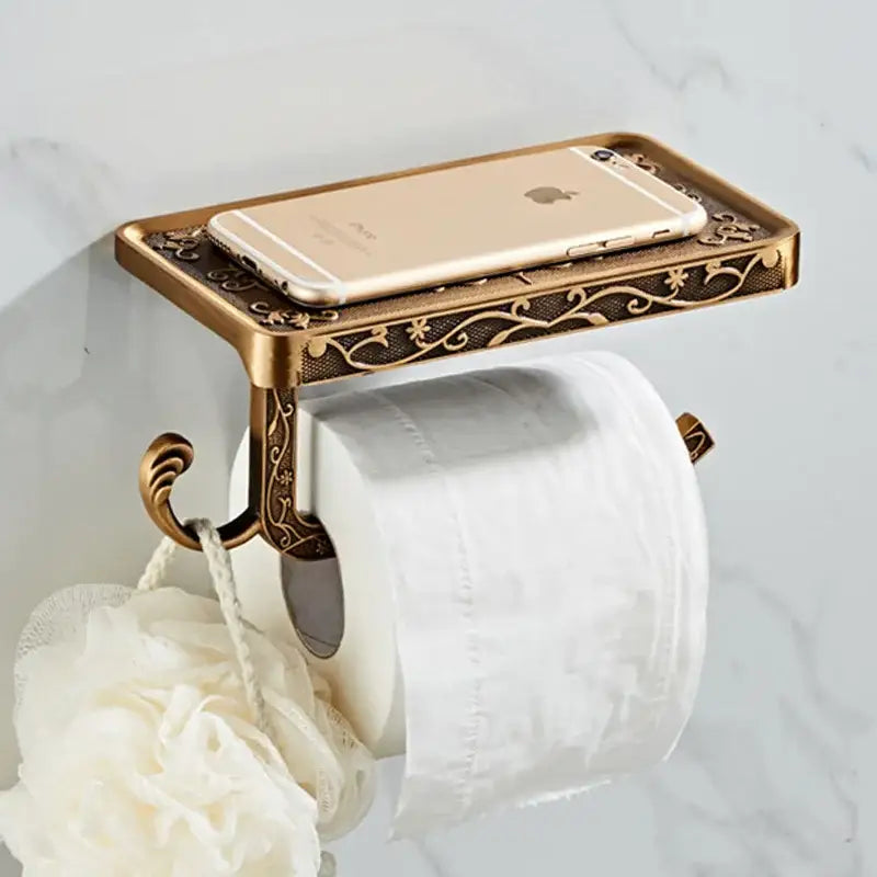 Antique brass toilet paper holder with ornate floral engraving and integrated phone rack holding a gold iPhone