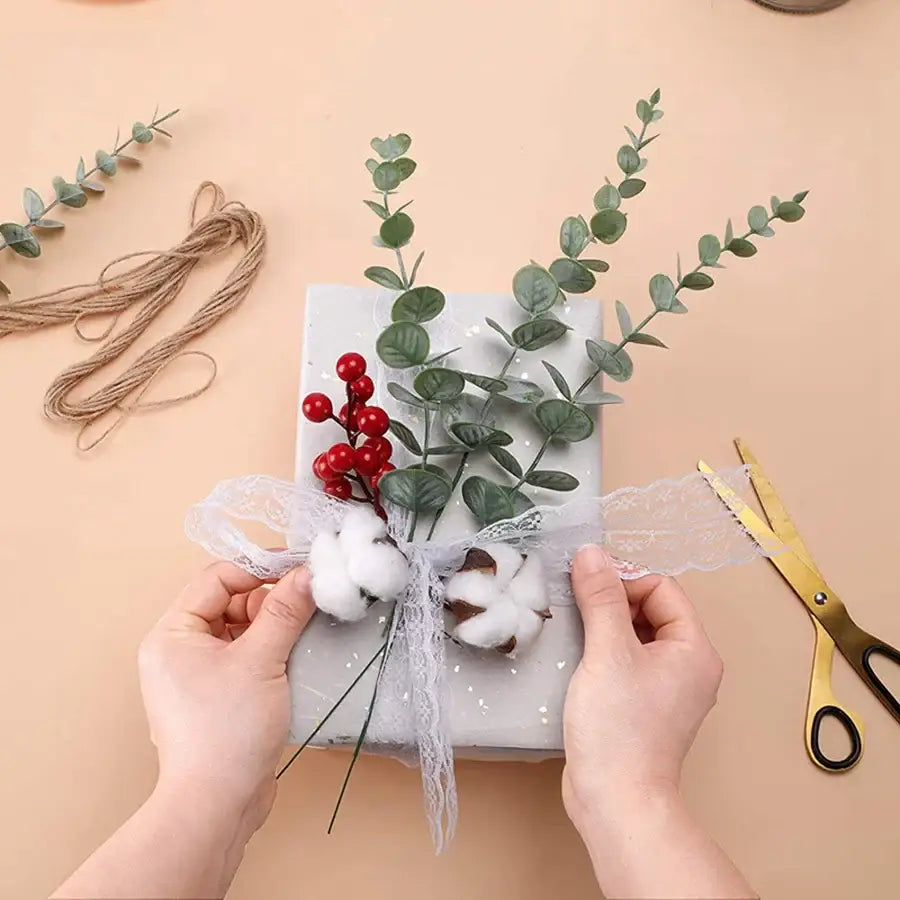 A gift wrapped in silver foil with a white lace bow and red berries decorated with artificial eucalyptus leaves by decorbites
