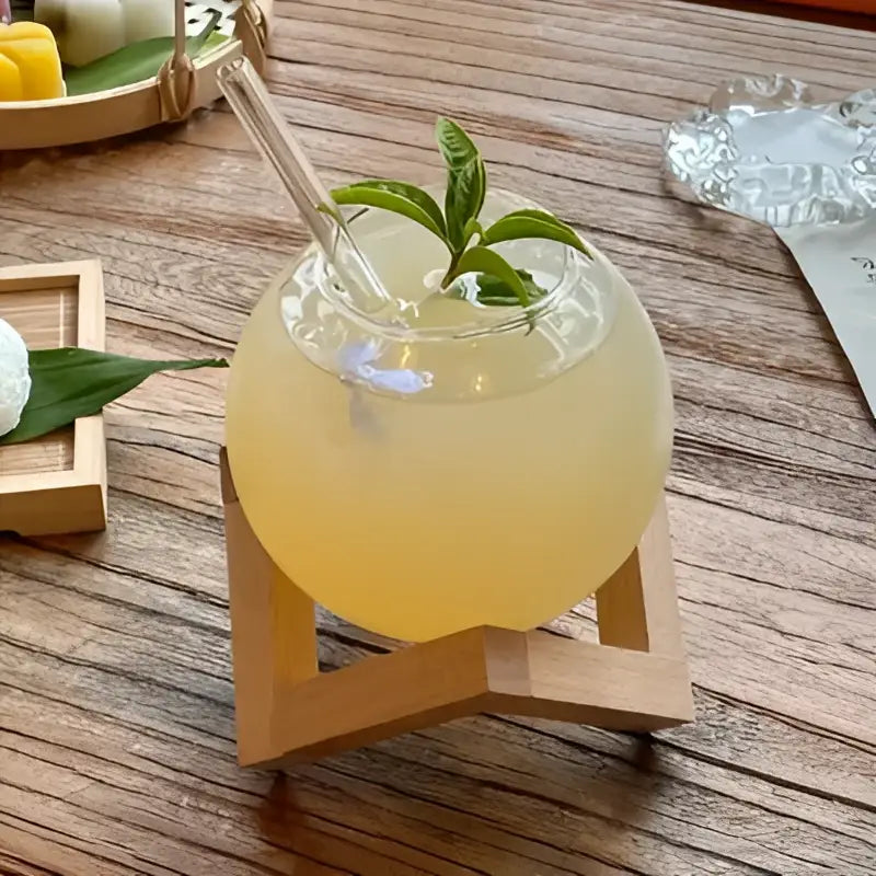 A clear glass ball shape coffee juice cocktail glass with pale yellow drink and fresh mint on a wooden stand