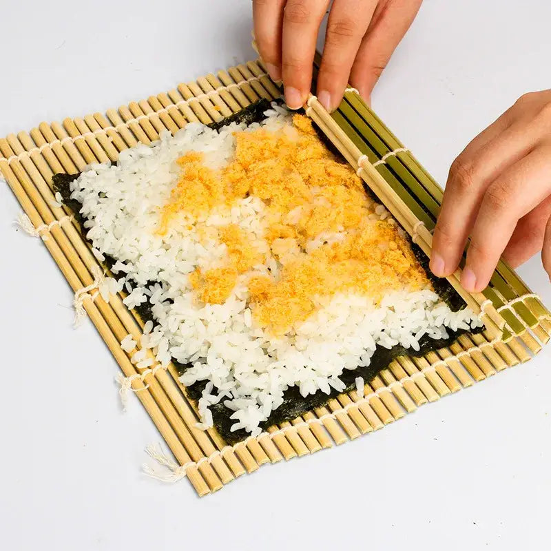 Bamboo sushi rolling mat with dark green nori sheet, white rice, and orange fish flakes for sushi rolling and onigiri rice paddles