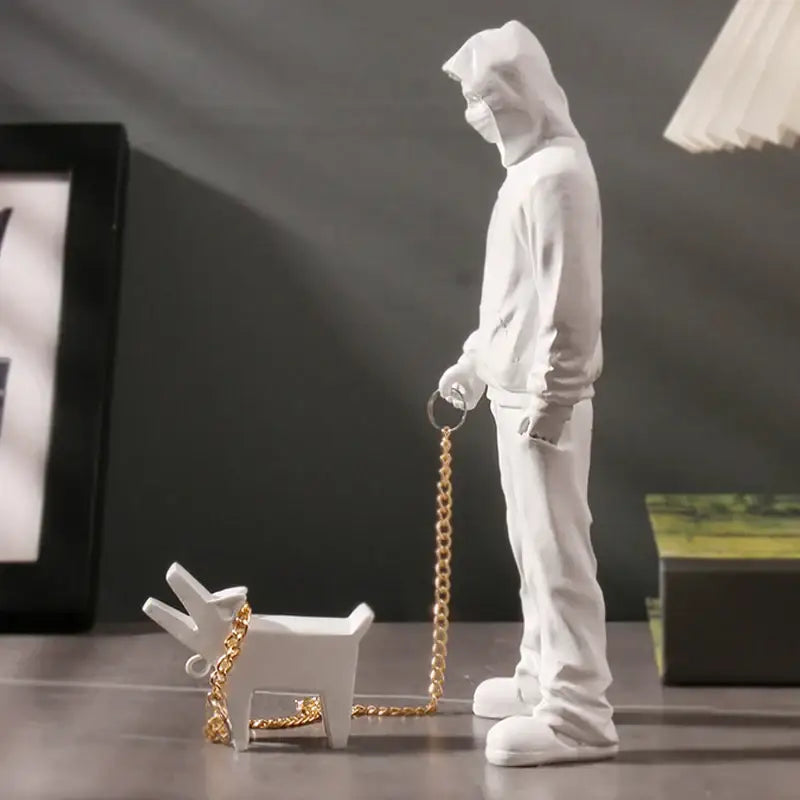 White resin banksy dog walker statue featuring a hooded figure holding a gold chain leash attached to a small dog object