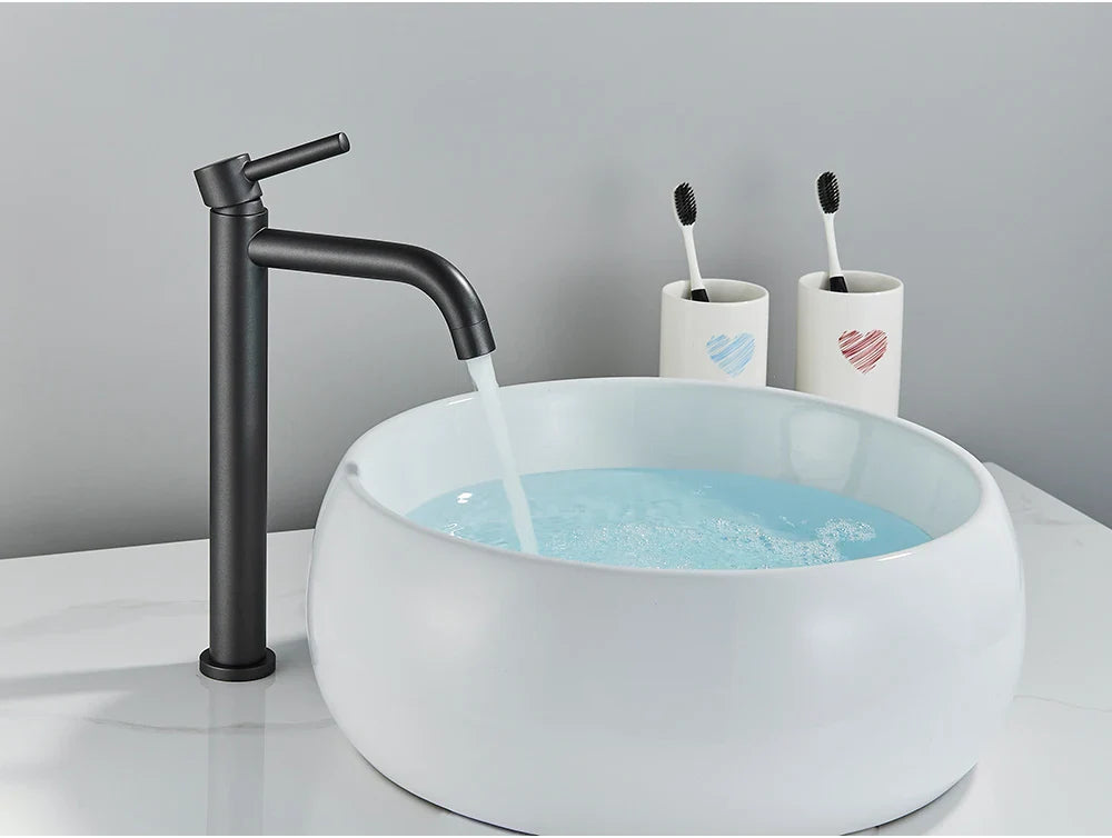 Black matte finish tall bathroom faucet with curved spout flowing water into white ceramic vessel sink