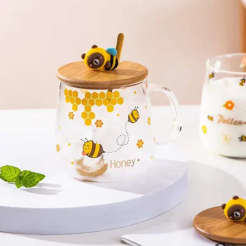Clear glass bee glass spoon milk cup with yellow bee illustrations and bamboo lid topped by bear bee figurine