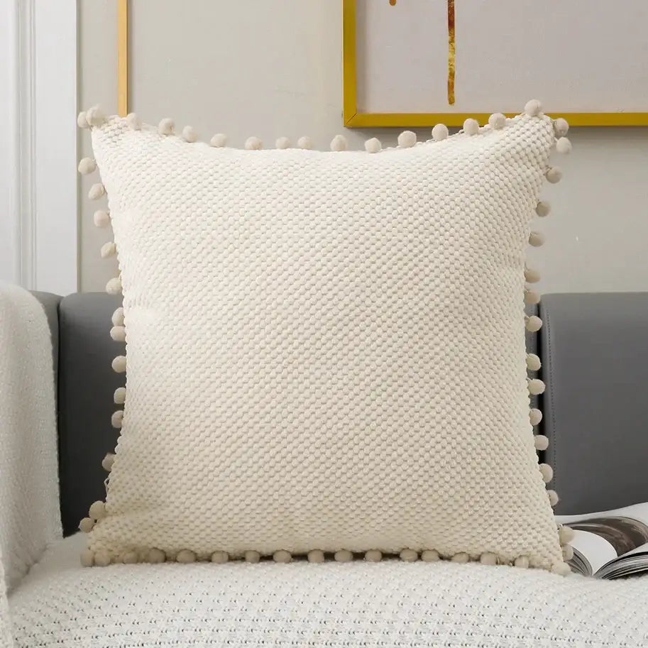 Cream-colored knit decorative pillow cover with textured woven pattern and soft pom-pom trim for living room decor