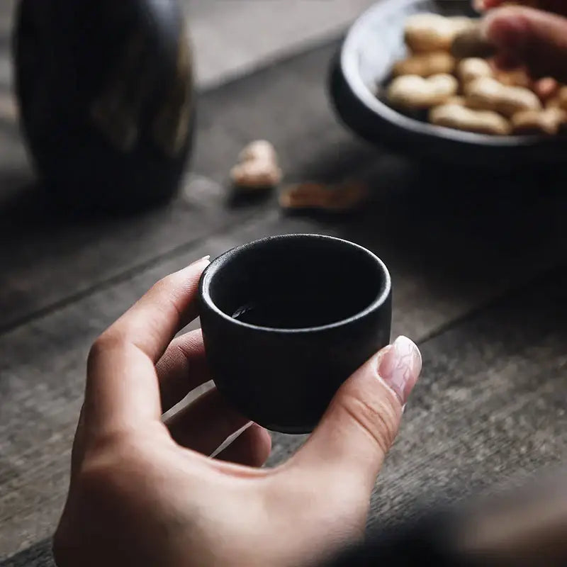 A hand holding a small matte black ceramic sake cup from the black and gold brushstroke ceramic sake liquor dispenser set