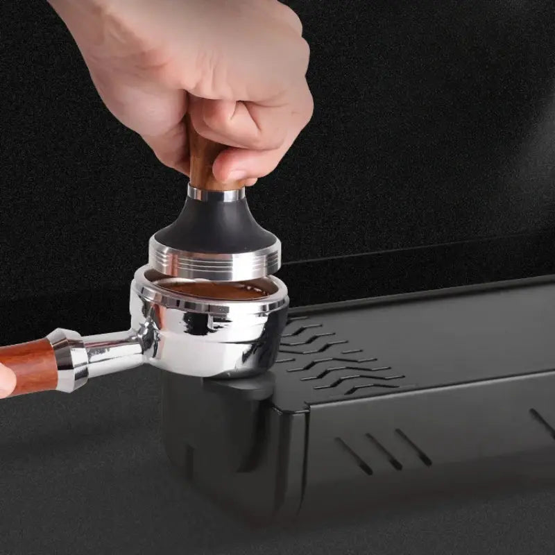A hand presses a wooden-handled silver coffee tamper into ground coffee on a black and green barista tamping station tray