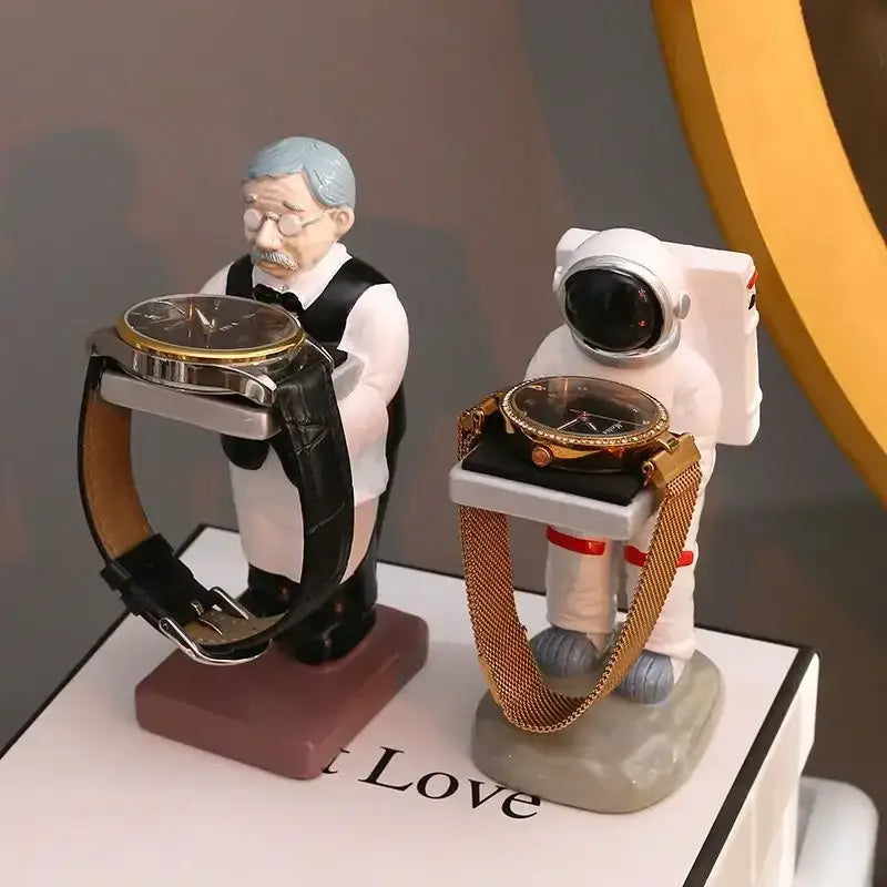 Two whimsical watch holders shaped like a vintage waiter and astronaut beside a black and iridescent purple cartoon bear watch stand bedroom decor