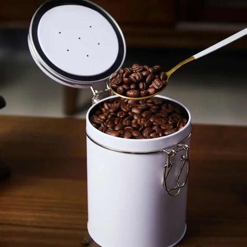 White cylindrical metal coffee canister with silver lid and clasp filled with dark roasted beans for hermetic food storage