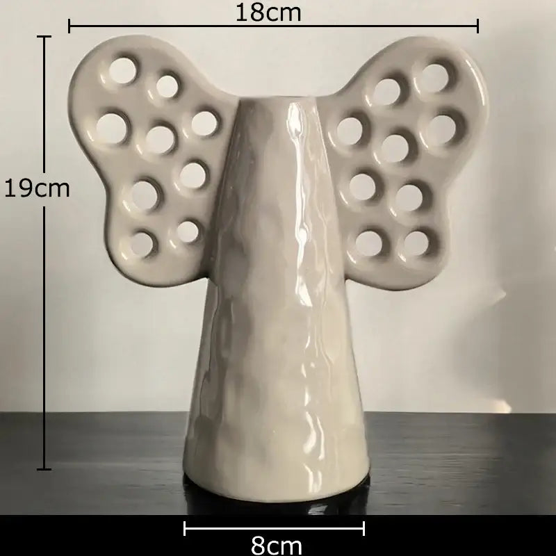 A glossy off-white ceramic angel figurine with butterfly wings next to a decorbites elephant ceramic vase with artificial flowers