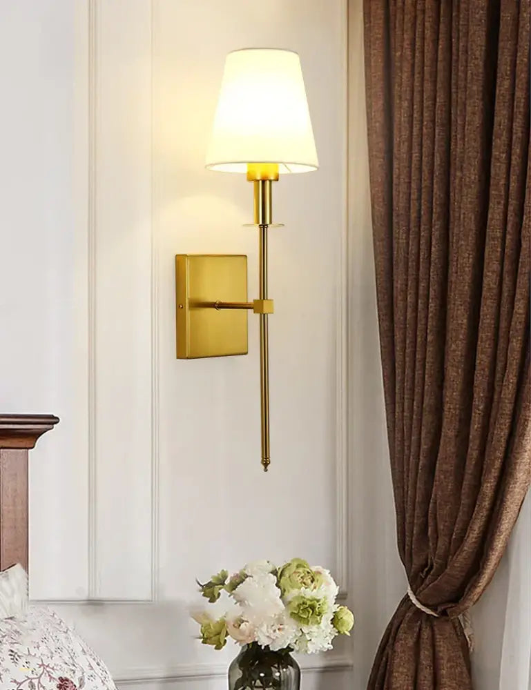 Gold-toned wall sconce with tapered off-white fabric shade featuring black brass accents for bedroom lighting
