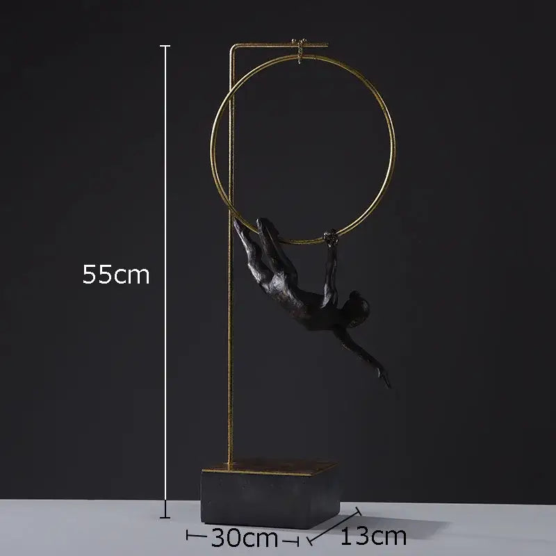A black and gold gymnastic acrobat resin sculpture with a sleek figurine hanging from a golden hoop on a stand