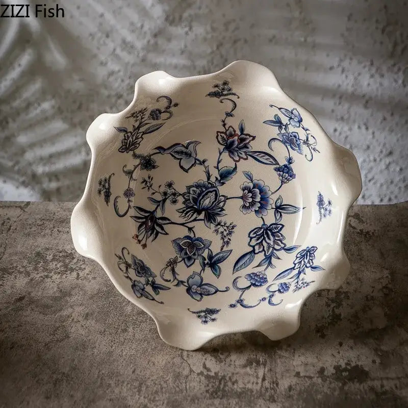 A cream-colored ceramic fruit bowl with scalloped edge and intricate blue flower pattern footed design on gold base
