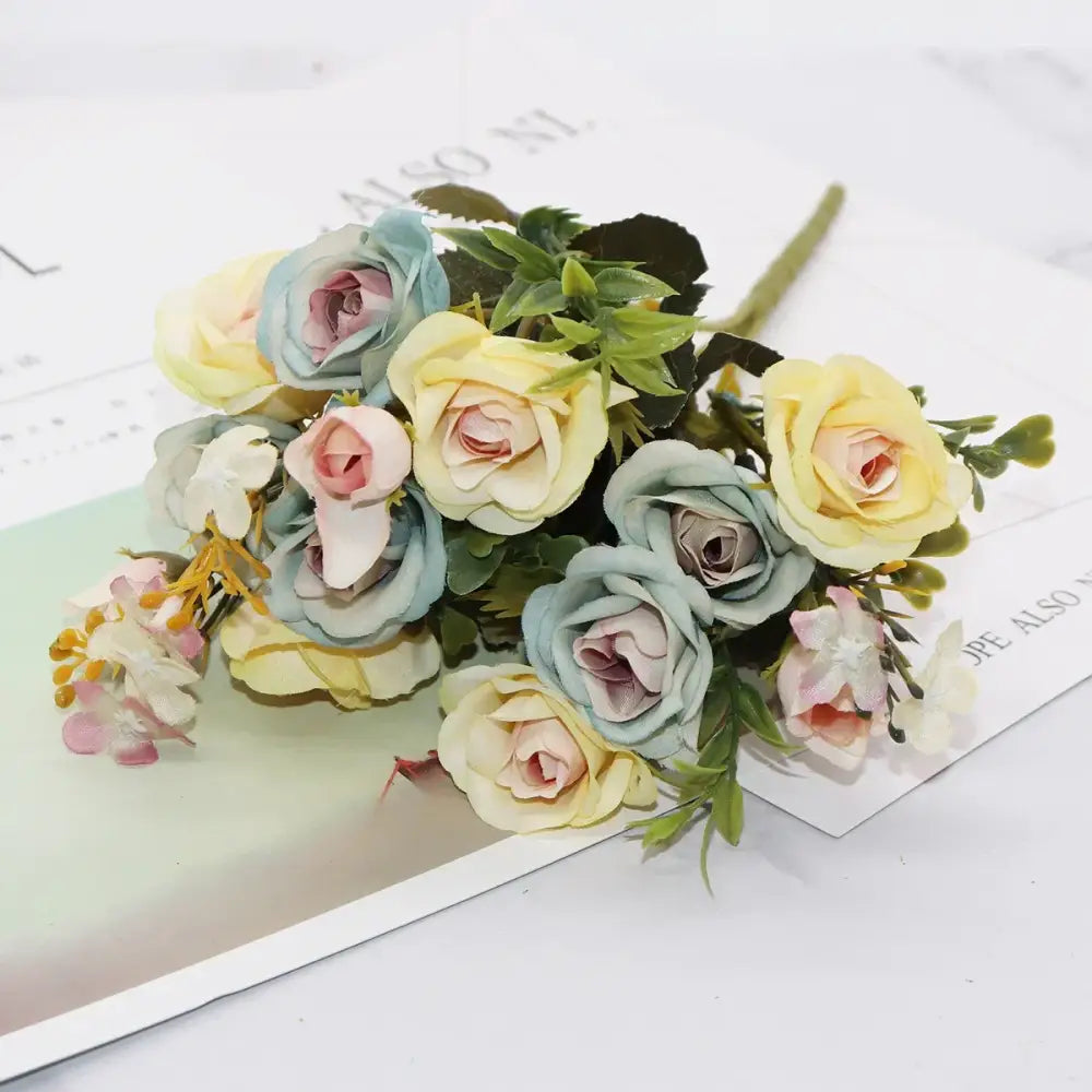A delicate bouquet of silk fake flowers featuring blue peony and tea rose in pastel yellow blush pink and green foliage