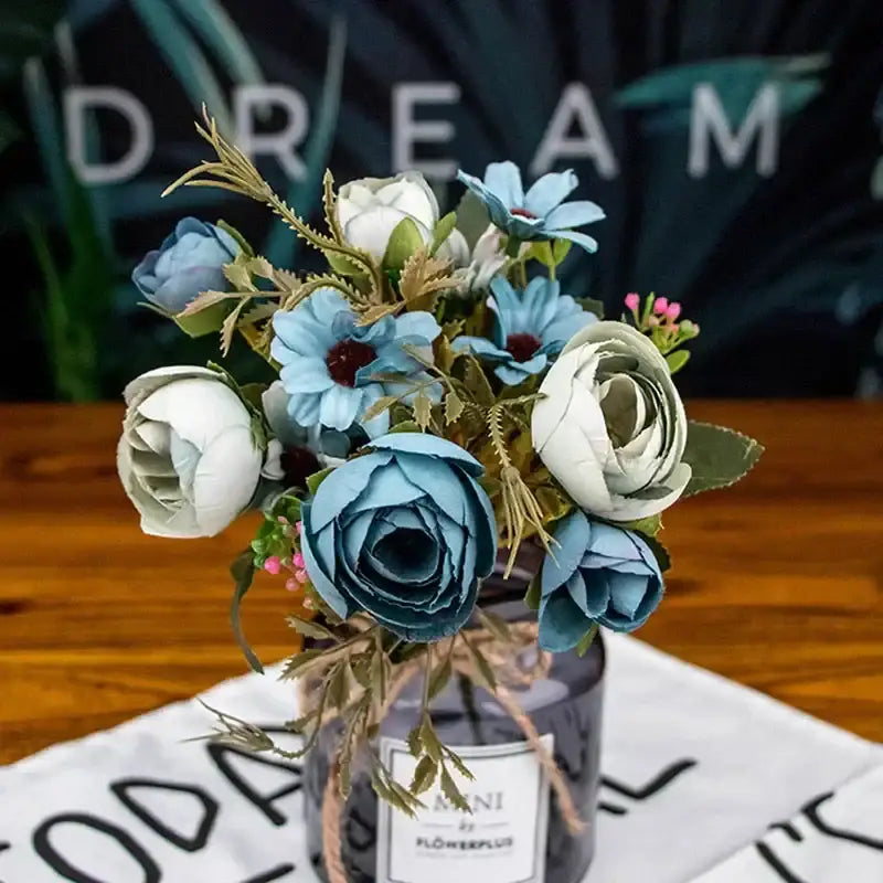 A delicate bouquet of silk fake flowers featuring blue peony and tea rose in a dark gray glass jar with twine wrap